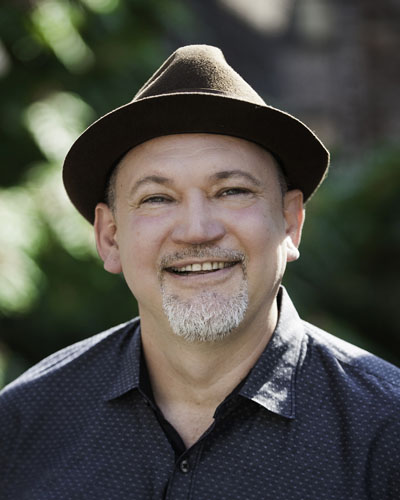 Un hombre latino blanco con una perilla corta y gris, con un sombrero fedora marrón y una camisa gris abotonada. Crédito de la foto: Michael Palma.