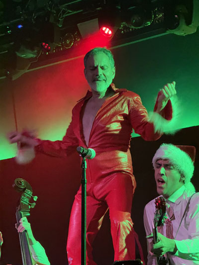 El Vez, un apuesto hombre chicano mayor, baila sobre un contrabajo vistiendo un mono de vinilo rojo con ribete de piel.