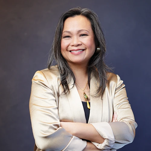 Smiling woman with long hair in a champagne blazer, arms crossed, in front of a dark blue background.