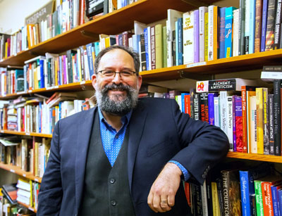 Michael es un hombre de cabello negro, barba negra y gafas que sonríe mientras se apoya en una estantería.