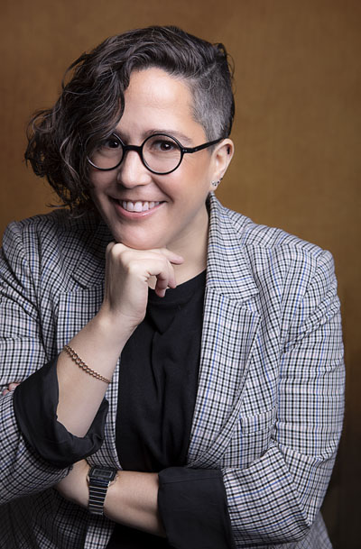 Una mujer con gafas negras redondas, cabello corto con un lado rapado y el otro rizado, una sonrisa y un atuendo compuesto por un blazer a cuadros y una camisa negra. Crédito de la foto: Odette Villareal
